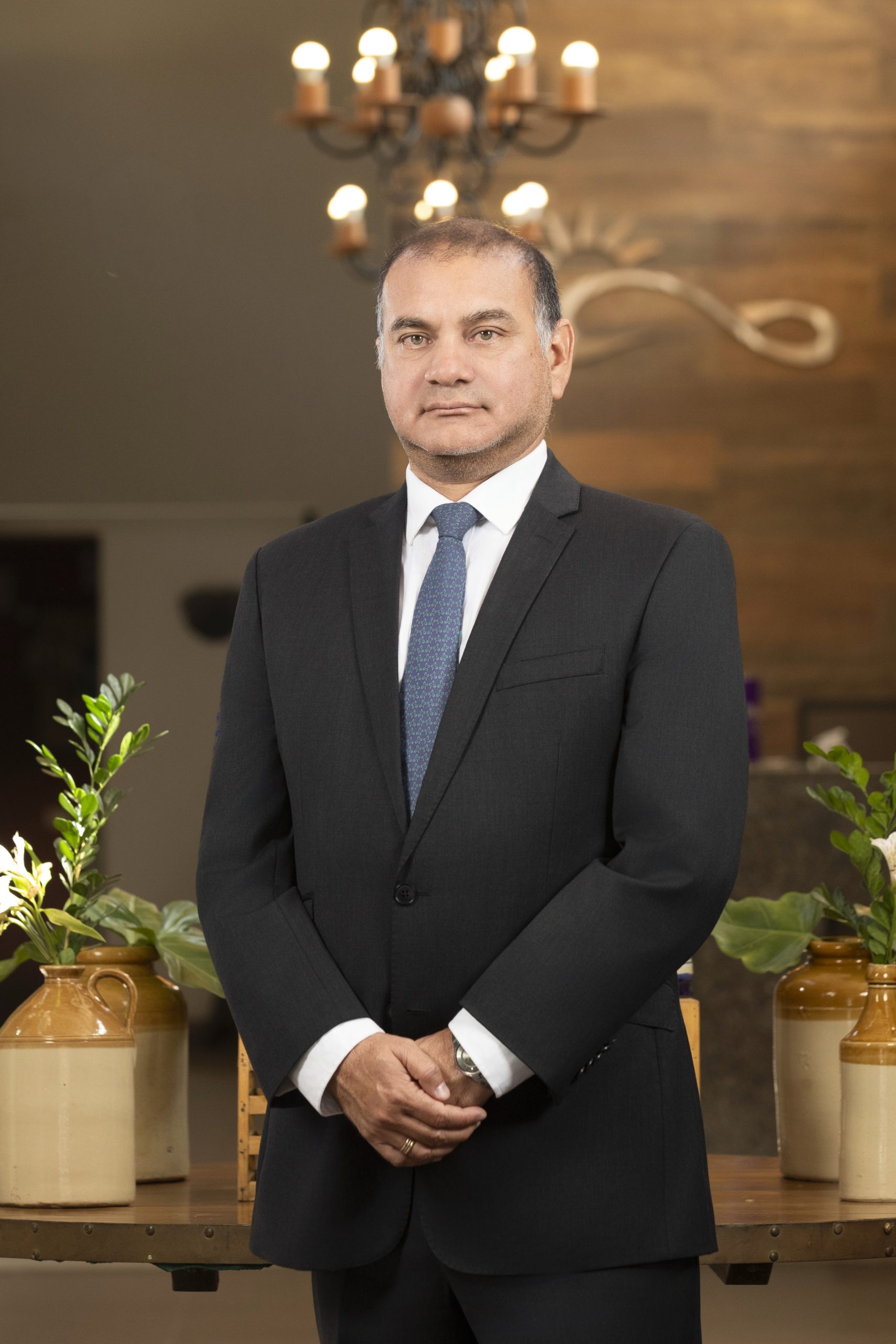 Miembros del Directorio 4 Retrato de Alejandro Molinari, ejecutivo de negocios de Caja Ica, de pie, con traje y corbata, en un entorno interior elegante con plantas y lámpara de fondo.